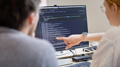 To personer, der arbejder sammen ved en computer, hvor den ene peger på koden på skærmen. Koden vises i et programmeringsmiljø med mørk baggrund.